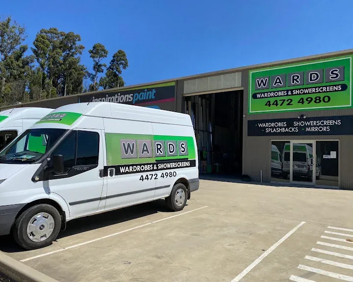 Two white Wards Wardrobes & Showerscreens vans parked outside the Wards workshop in Batemans Bay, under a green sign listing wardrobes, showerscreens, splashbacks and mirrors and the phone number 4472 4980.